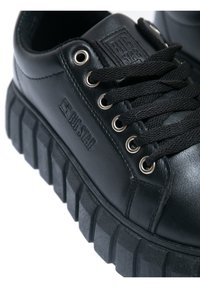 Mustad mustad sneakerid tekstuurses kummitallaga, millel on metallist aukude ja mustade paeltega. Peal on graveeritud logo brändimiseks.