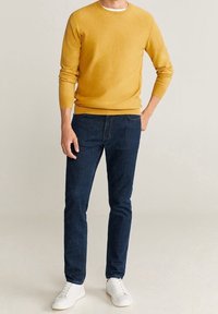 Muž v hořčicově žlutém svetru, tmavě modrých džínách a bílých teniskách stojí s jednou rukou v kapse na neutrálním pozadí.