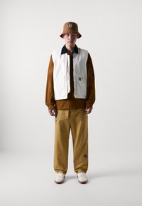 Brauner Bucket Hat, weiße Zip-Up-Weste, braunes Langarmhemd, gelbe weite Hose mit Stickerei und weiße Sneaker mit braunen Sohlen.