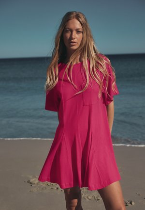 Jeune femme aux longs cheveux blonds portant une robe ample rose vif, debout sur une plage de sable avec un océan calme en arrière-plan.