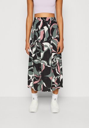 Femme portant une jupe midi fluide taille haute avec des motifs floraux abstraits en noir, blanc, rose et vert, associée à des baskets blanches et des chaussettes.