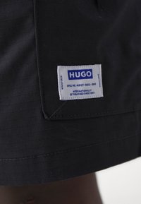 Tissu noir avec une étiquette de marque rectangulaire blanche HUGO cousue près de l’ourlet, avec un texte bleu et de petits détails imprimés.
