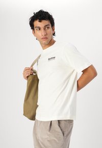 Bílá bavlněná tričko s krátkým rukávem a logem Barbour, spárované se světle béžovými kordovými kalhotami a držící hnědou plátěnou tašku.