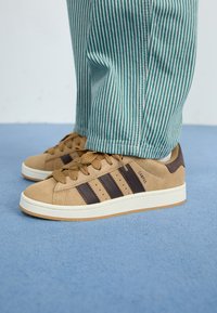 Brown suede sneakers with three dark brown stripes, white sole, and beige laces. Paired with teal and white striped pants.