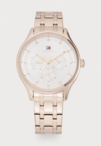 Reloj metálico de oro rosa con una esfera circular blanca, tres sub-esferas, marcadores dorados y una pulsera de estilo eslabonada. Presenta el logo de la marca.