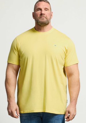 Brodaty mężczyzna stojący, ubrany w jednolity żółty T-shirt z krótkim rękawem i niebieskie dżinsy na neutralnym tle.