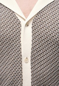 Gros plan d'une personne portant un cardigan crème avec un motif zigzag noir et beige et deux boutons visibles autour de l'encolure.