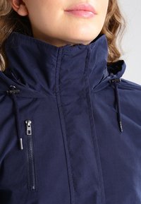 Granatowa kurtka z wysokim kołnierzem, regulowanymi sznurkami i przednią kieszenią na zamek. Gładki materiał o czystym, dopasowanym kroju.