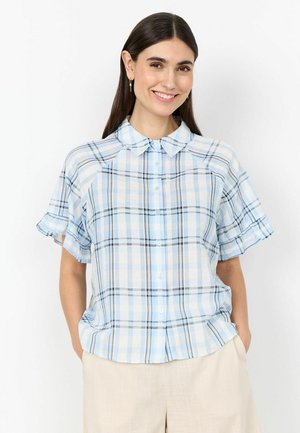 Donna che sorride indossando una camicetta a maniche corte a quadri blu e bianchi con colletto e pantaloni beige, con le mani nelle tasche.