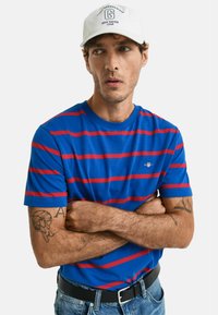 Blauw gestreept T-shirt met horizontale rode strepen, gemaakt van katoen. Gecombineerd met een witte pet met een logo en een zwarte riem. Denim jeans.