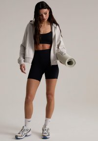 Sudadera gris claro con cremallera, sujetador deportivo negro y pantalones cortos de cintura alta negros; sosteniendo una esterilla de yoga enrollada; zapatillas blancas y azules con acentos.