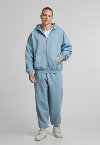 Svetlomodrá mikina na zips s veľkými prednými vreckami, skombinovaná s nohavicami v rovnakom odtieni. Vyrobená z mäkkej látky, s rebrovanými manžetami a lemom. Biele tenisky.