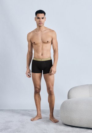 Tânăr stând desculț pe un podea cu covor, purtând chiloți boxer negri cu talie aurie, lângă o canapea curbată de culoare crem.