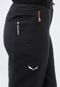 Μαύρο ελαστικό παντελόνι με τσέπη με φερμουάρ, πορτοκαλί τράβηγμα φερμουάρ, λεπτομέρειες Durastretch και λευκό λογότυπο στον μηρό.