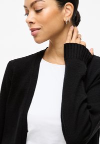 Μαύρος κάρντιγκαν με χαλαρή εφαρμογή, ριμπ μανσέτες και ανοιχτό μπροστινό σχέδιο, φορεμένος πάνω από ένα απλό λευκό πουκάμισο. Ορατή μαλακή υφή.