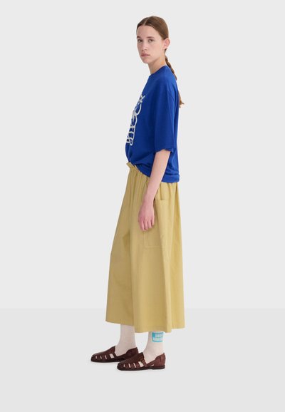 Femme aux cheveux bruns tressés, portant un t-shirt bleu avec un motif graphique, une jupe large beige, des chaussettes blanches avec du texte bleu, et des sandales marron, debout de profil.