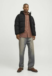 Sort dunjakke med hætte over en brunternet skjorte. Lyseblå jeans og brune sko fuldender det afslappede look.