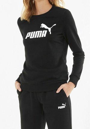Kobieta w czarnej bluzie Puma i pasujących spodniach z białymi logo Puma, ręce w kieszeniach, na jednolitym tle.