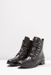 Les bottines en cuir noir présentent une finition lisse, un design à bout rond, des œillets argentés brillants avec une fermeture à lacets et un talon bas.