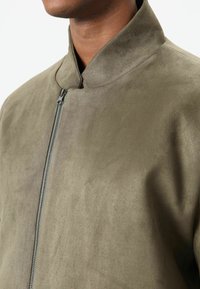 Olivgrüne Wildlederjacke mit hohem Kragen, Frontreißverschluss und weicher Textur. Nahtdetails verleihen dem minimalistischen Design subtile Struktur.