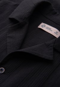 Gros plan d’une chemise noire texturée à manches courtes avec col, boutons noirs et une étiquette de marque visible à l’intérieur du col.