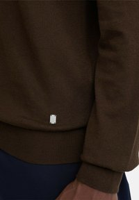 Bruine gebreide trui met geribbelde manchetten en zoom. Heeft een klein zilveren logolabel vlak bij de onderkant. Gladde textuur met een aangesloten pasvorm.