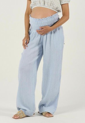 Femme enceinte portant un pantalon de maternité ample bleu clair à jambes larges, tenant son ventre, assorti d'un haut court à carreaux et de sandales.