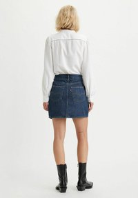 Jupe en denim mini de couleur bleu foncé avec une coupe structurée, des poches arrière et une taille haute. Associée à un blouse blanche à manches longues et des bottines noires.