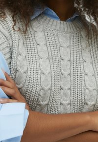 Jasnoszary sweter z dzianiny z wzorem w warkocze, okrągłym dekoltem i prążkowanym wykończeniem. Nosi się go na niebieskiej koszuli z kołnierzykiem.