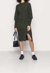 Robe tricotée vert foncé avec texture côtelée, manches longues bouffantes et fente latérale. Sac à main noir structuré avec finition brillante. Chaussures noires et chaussettes blanches.