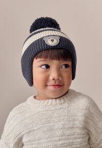 Strickmütze in dunkelgrau und cremefarbenen Streifen, mit einer runden Bommel oben und einem Bärenaufnäher auf der Vorderseite. Weiche, gerippte Textur.