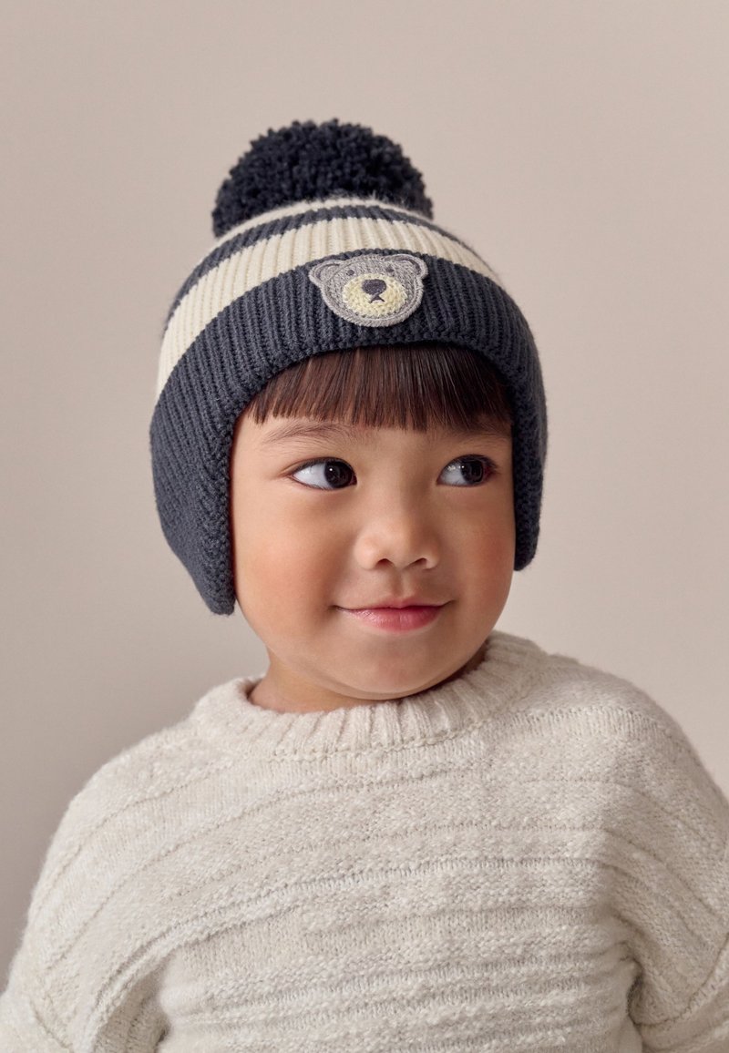 Strickmütze in dunkelgrau und cremefarbenen Streifen, mit einer runden Bommel oben und einem Bärenaufnäher auf der Vorderseite. Weiche, gerippte Textur.