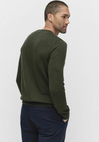 Tummanvihreä pitkähihaisen neuleen, jossa on pyöreä pääntie, ribbaatut hihansuut ja helma, käytetään tummansinisien housujen kanssa, nähtynä takaa. Pehmeä rakenne ja istuva muoto.