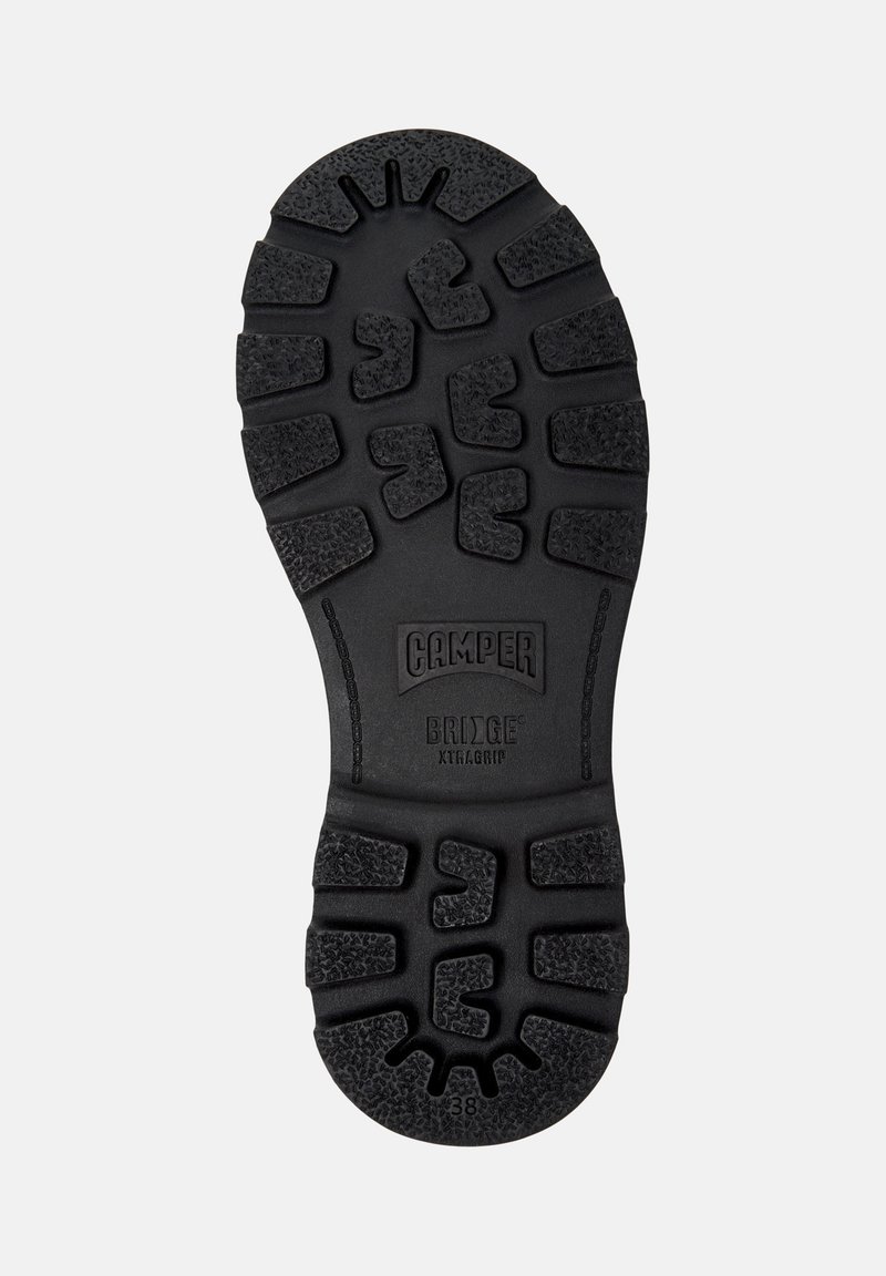 Zwarte rubberen schoenzool met diepe profielen, merknnaam "Camper," model "Bridge XTRAGRIP," en maat "38" zichtbaar op de onderkant.