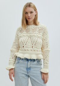 Beigefarbener gestrickter Pullover mit Spitzenmuster, gerippten Bündchen und einem Peplum-Saum. Kombiniert mit hellblauen Jeans.