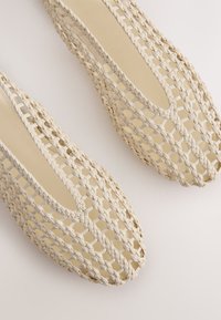 Chaussures plates beiges tissées avec un design en treillis ouvert et des bouts carrés arrondis sur un fond clair.