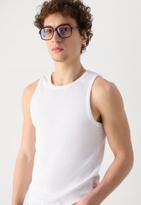 Un débardeur blanc côtelé avec un col rond, porté par une personne aux cheveux frisés, associé à des lunettes rectangulaires marron avec des verres bleus.