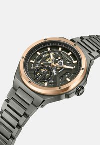 Automatische horloge met een zwarte skeleton-dial, gouden accenten, een roestvrijstalen band en een gestructureerde grijze kast. Beschikt over een uniek ontwerp met zichtbare mechanica.