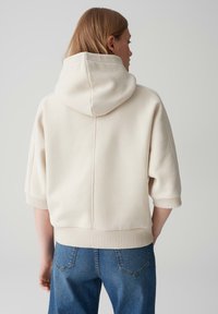 Crème hoodie met driekwartmouwen en geribbelde zoom. Gemaakt van zacht materiaal, met een naaddetail aan de achterkant. Gecombineerd met een blauwe jeans.
