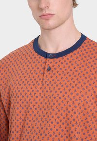 Maglietta arancione a maniche corte con colletto blu navy, caratterizzata da un motivo ripetitivo di ancore e raggi di sole. Chiusura a due bottoni sul collo.