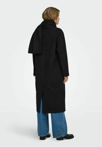 Cappotto di lana nera con una vestibilità rilassata, colletto a revers e spacco sul retro. Abbinato a jeans blu a gamba larga e stivaletti neri.