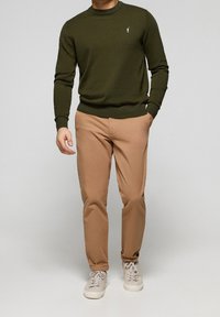 Mörkgrön stickad tröja med rund hals, matchad med tan chinos och ljusgrå sneakers. Anmärkningsvärd logotyp på tröjans bröst.