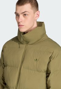 Olive zaļš pūkains mētelis ar augstu apkakli, ar rāvējslēdzēju priekšā un smalku logo detaļu uz krūtīm. Tekstūrēts, viegls materiāls.