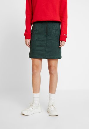 Groene suede mini-jurk met een ritssluiting aan de voorkant, gecombineerd met een rode sweatshirt en witte sneakers, voorzien van witte sokken tot de enkels.