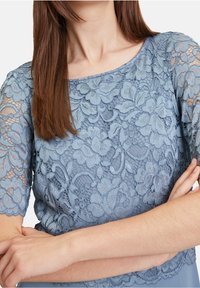 Kvinna i en blå spetsblus med blommönster, med armarna i kors, långt brunt hår och neutral hudton.