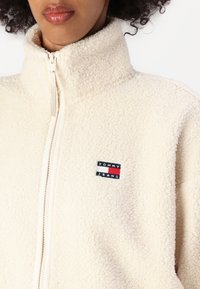 Krémová fleecová bunda s vysokým golierom a zipsom. Obsahuje malé modré, červené a biele logo "TOMMY JEANS" na ľavej strane.