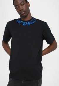 Svart kortärmad T-shirt med blå broderad text runt halsringningen, tillverkad av mjukt material, med en avslappnad passform och en diskret sidologo. 