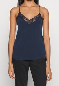 Žena v tmavě modrém topu na ramínka s krajkovým lemem a tenkými ramínky, v kombinaci s černými kalhotami, na jednobarevném světlém pozadí.