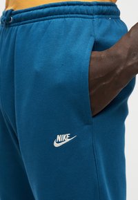 Μπλε φόρμες Nike με ελαστική μέση, πλαϊνές τσέπες και λευκό λογότυπο στο αριστερό μηρό. Μαλακή υφή, καθημερινό σχέδιο.