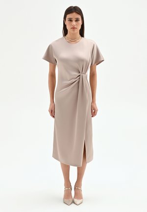 Donna in piedi che indossa un abito midi beige con maniche corte, dettaglio con nodo laterale, spacco, collana d'oro e sandali con cavigliera dorata.